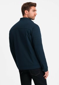 Veste bleu marine en tissu texturé, col montant et détails de couture horizontale sur les épaules. Design épuré avec une silhouette ajustée.