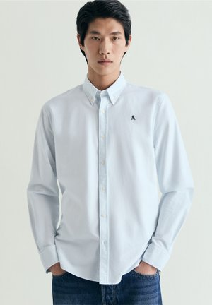 Tânăr care poartă o cămașă albastru deschis cu nasturi, având un mic emblemă neagră cu craniu și oase încrucișate pe piept, cu mâinile în buzunarele blugilor albaștri.