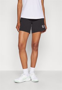  svarte treningsshorts med en tettsittende design og hvit "EA"-logo på venstre ben. Kombinert med en hvit skjorte og hvit-grønne sneakers.