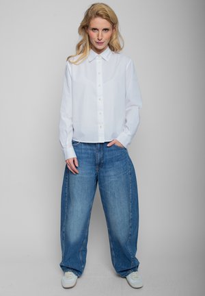 Blonde Frau steht frontal, trägt ein weißes, geknöpftes Hemd, lockere blaue Jeans und weiße Turnschuhe vor einem schlichten grauen Hintergrund.