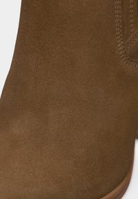 Καφέ μποτάκι από σουέτ με ορατές ραφές και λεία υφή, παρουσιάζοντας το μπροστινό και το πλάγιο μέρος πάνω σε λευκό φόντο.