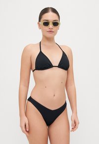 Juodas bikini maudymosi kostiumėlis su trikampiu viršumi, plonomis juostelėmis ir raukšlėta tekstūra. Modelis, vilkintis žalius akinius nuo saulės, stovi prieš paprastą foną.