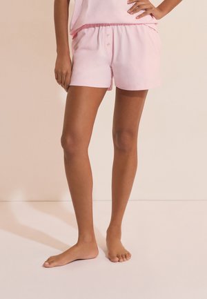 Femme portant un short à boutons de couleur rose clair et un haut sans manches assorti, debout pieds nus sur un sol pâle.