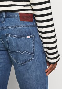 Jeans de mezclilla azul con un diseño distintivo en el bolsillo trasero que presenta un patrón en zigzag y una etiqueta de logo rojo, combinados con un suéter a rayas en blanco y negro.