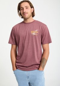 Bordeauxrotes Baumwoll-T-Shirt mit kurzen Ärmeln und Rundhalsausschnitt, featuring ein gelbes Grafiklogo auf der linken Brust. Kombiniert mit hellblauen Jeans.