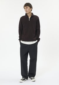 Dunkelbrauner Pullover mit einem halben Reißverschluss-Kragen, über einem weißen Hemd getragen. Kombiniert mit lockeren schwarzen Hosen und schwarzen Turnschuhen. Einfaches Design, lässiger Look.