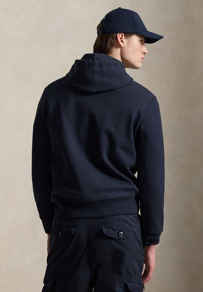 Jonge man die een marineblauwe hoodie, marineblauwe cargobroek en een marineblauwe baseballpet draagt, lichtjes opzij gedraaid tegen een neutrale achtergrond.