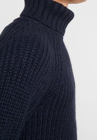 Gros plan sur une personne portant un pull à col roulé épais en tricot bleu foncé avec un motif texturé.