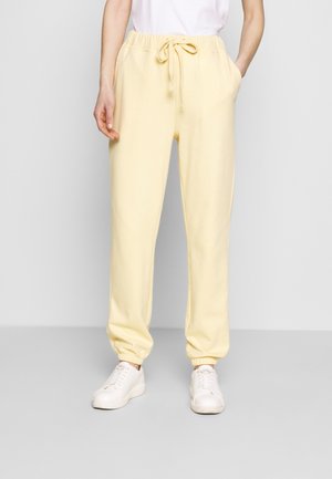 Personne portant un pantalon de jogging jaune pâle à cordon et des baskets blanches, debout devant un fond clair uni.