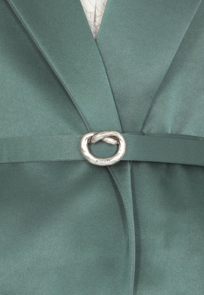 Blazer din satin verde cu detaliu în formă de cataramă circulară argintie la talie și rever decupat. Textură netedă cu elemente de design structurat.