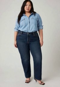 Svetlo modra srajca z gumbi in dvema žepoma na prsih, v kombinaciji s temno modrimi visokimi jeans hlačami. Model nosi črne, zavezane sandale s peto.