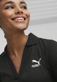 Svart ribbet polo-skjorte med v-hals, preget av en hvit brodert Puma-logo på venstre side. Glatt tekstur og tett passform.