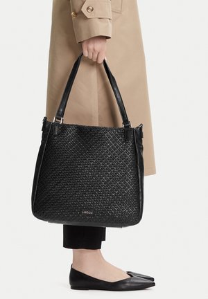 Ruka držící velkou černou pletenou tašku s dvojitými uchy, viditelný béžový kabát, černé kalhoty a černé špičaté baleríny.