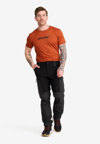 Man med tatueringar iklädd en orange RVRC t-shirt, svarta cargobyxor och svarta vandringsskor som går med händerna i fickorna.