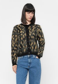 Mulher com um cardigã felpudo de padrão leopardo preto e castanho, vestindo jeans azuis, com uma mão no bolso, contra um fundo claro e simples.