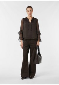 Zwarte doorschijnende blouse met gerimpelde mouwen, V-hals, gecombineerd met donkerbruine uitlopende broek. Model houdt een bruine shopper vast.