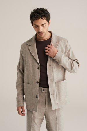 Svetlo rjava volnena jakna z izrezanim ovratnikom, dvema velikima žepoma in temnimi gumbi. Oblečena preko rjave srajce.