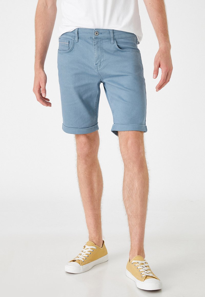 Pantalones cortos de mezclilla azul claro con puños enrollados, diseño de cinco bolsillos y cierre de botón. Llevados con zapatillas amarillas y una camiseta blanca.