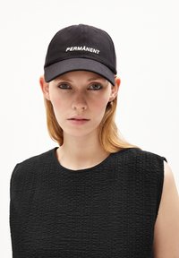 Black cap with "PERMANENT" embroidered in white. The wearer has straight blonde hair, wearing a textured black top with a round neckline.