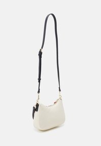 Kleine crèmekleurige schoudertas met een lange verstelbare zwarte riem en hardware in goudkleur, getoond tegen een witte achtergrond.