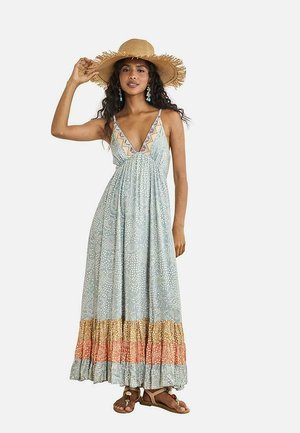 Mujer con un vestido largo sin mangas estampado, sombrero de paja de ala ancha, pendientes turquesa y sandalias, posando con una mano sobre su sombrero.