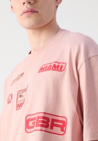 T-shirt en coton rose avec divers imprimés graphiques rouges, comprenant du texte et des motifs, avec un col rond et des manches courtes.