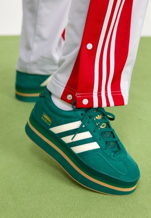 Zielone sneakersy Adidas Gazelle z białymi paskami noszone z białymi i czerwonymi spodniami dresowymi na zielonej podłodze.