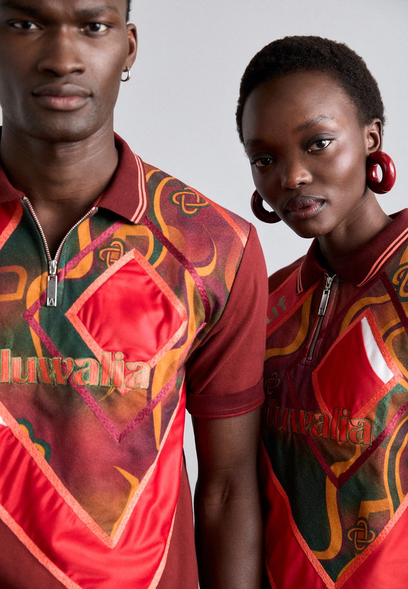 Twee personen dragen poloshirts met korte mouwen, versierd met levendige, gestructureerde patronen in rood, groen en geel, met ritskraag en geborduurde logo's.