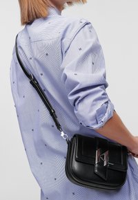 Borsa a tracolla in pelle nera con una texture liscia, dotata di un accento in logo argento e tracolla regolabile, indossata sopra una camicia a righe blu.