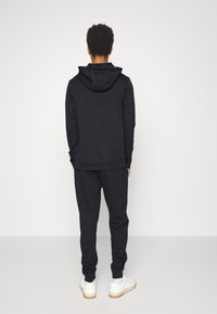 Svart hettegenser med kengurulomme, matchende svarte joggebukser, ribbestrikkede mansjetter og hem. Hvite sneakers med gummisåler. Enslig bakgrunn.