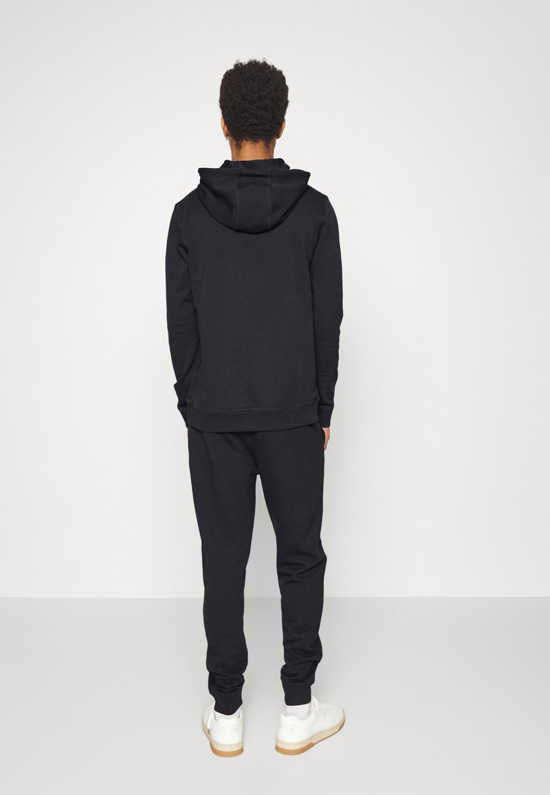 Svart hettegenser med kengurulomme, matchende svarte joggebukser, ribbestrikkede mansjetter og hem. Hvite sneakers med gummisåler. Enslig bakgrunn.