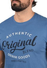 Modra bombažna majica z okroglim izrezom, kratkimi rokavi in natisnjenim besedilom v beli in mornarsko modri barvi, ki pravi "Avtentična Originalna Mešanica Denim Blaga."
