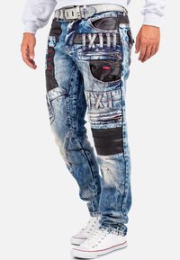 Pantalons cargo en denim avec un délavage bleu, accents en cuir noir, plusieurs poches et coutures décoratives en rouge. Coupe décontractée, détails vibrants.