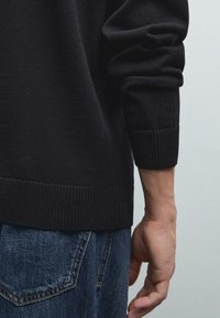 Schwarzer Strickpullover mit gerippten Bündchen, kombiniert mit blauen Jeans. Der Stoff hat eine glatte Textur und ein tailliertes Design.