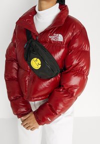 Czerwona kurtka puchowa o błyszczącym wykończeniu z logo, połączona z czarną nerka z żółtą buźką. Pod spodem biała golf.