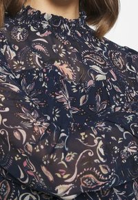 Blusa blu navy con motivi floreali in rosa e verde. Caratterizzata da un colletto arricciato e una texture trasparente, che conferisce un aspetto leggero.