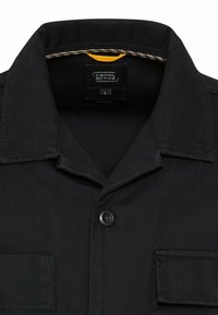 Czarna koszula overshirt z klasycznym kołnierzykiem, zapięciem na guziki z przodu oraz kieszenią na piersi. Metka wskazuje rozmiar L. Teksturowana tkanina i akcentujące przeszycia.