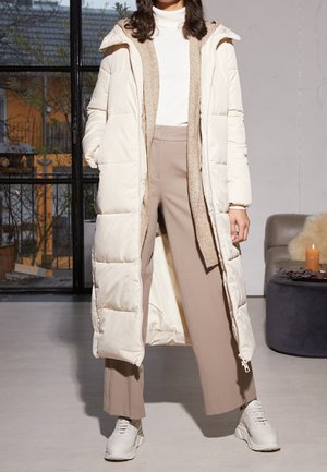 Lange gewatteerde jas in off-white, met een gestructureerde beige voering en capuchon. Gecombineerd met beige broeken en witte sneakers, in een lichte interieur.