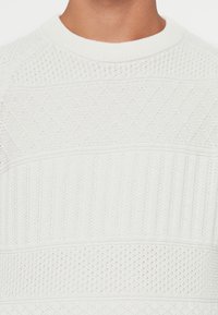 Hvid strikket sweater med teksturerede mønstre, vertikale og horisontale ribkanter, rund halsudskæring og åndbare, åbne væv-sektioner.