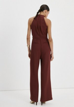 Frau mit geflochtenem Haar, die einen ärmellosen, bordeauxroten Overall mit hohem Kragen und beige Heels trägt, vor einem schlichten weißen Hintergrund stehend.