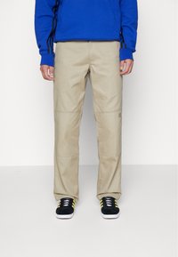 Pantalones beige con un corte relajado, que presentan detalles de costura y bolsillos laterales, combinados con zapatillas negras y una sudadera azul.