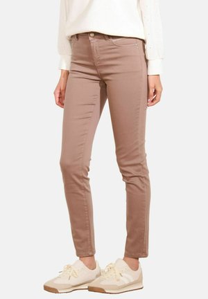 Donna che indossa pantaloni attillati color marrone chiaro, una blusa bianca e sneakers color crema in piedi contro uno sfondo neutro.