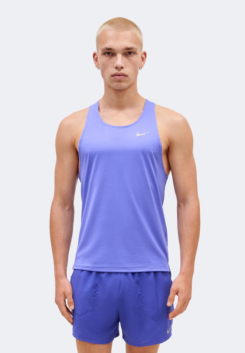 Νέος άνδρας με ξυρισμένο κοντό κούρεμα που φοράει ανοιχτό μωβ αμάνικη αθλητική μπλούζα και ταιριαστά σορτς, στέκεται μπροστά σε απλό φόντο.