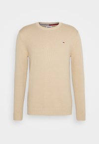 Pull tricoté beige avec poignets et ourlet côtelés. Il présente un col rond et un petit logo sur la poitrine. Texture douce et coupe standard.