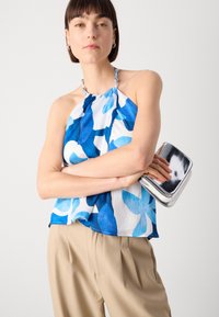 Haut halter bleu et blanc à motif avec un tissu texturé, associé à des bretelles en perles. Pochette en argent à surface lisse et légère brillance.