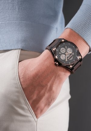 Muñeca con reloj negro de múltiples esferas de la marca Police, mano metida en el bolsillo de un pantalón claro, con la manga de un suéter azul claro visible.