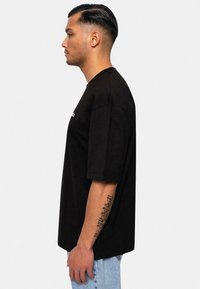 Sort t-shirt med korte ærmer lavet af bomuld, med rund hals og en løs pasform, set fra siden mod en lys baggrund.