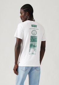 Mann med flettehår iført en hvit Levi's T-skjorte med grønne grafiske trykk og lyseblå jeans, vendt sidelengs mot en ensfarget bakgrunn.