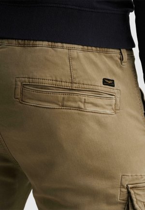 Primer plano de pantalones kaki con un bolsillo trasero de ojal y una pequeña etiqueta de marca rectangular negra cerca de la cintura, combinados con una camiseta negra de canalé.
