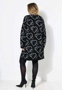 Knielanger schwarzer Pullover mit weißem Herzmuster, langen Ärmeln, lässiger Passform und weicher Textur. Getragen über schwarzen Strumpfhosen und High-Heel-Stiefeln.